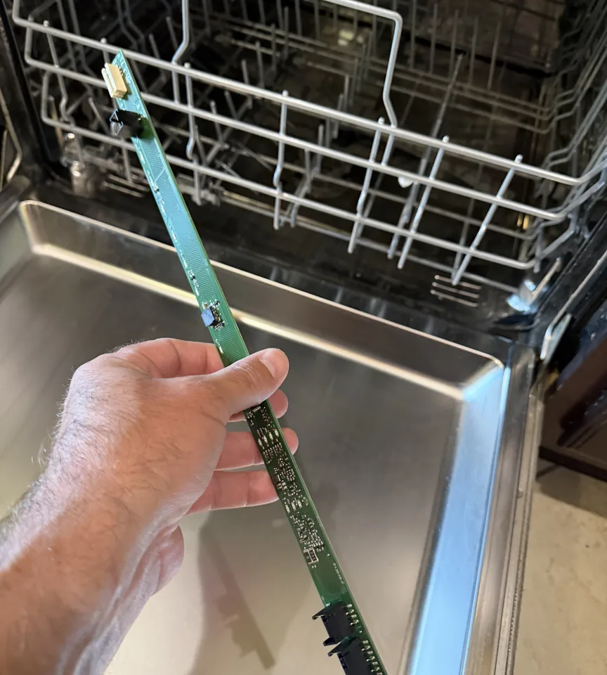 A long, thin dishwasher control board held up in front of an open dishwasher