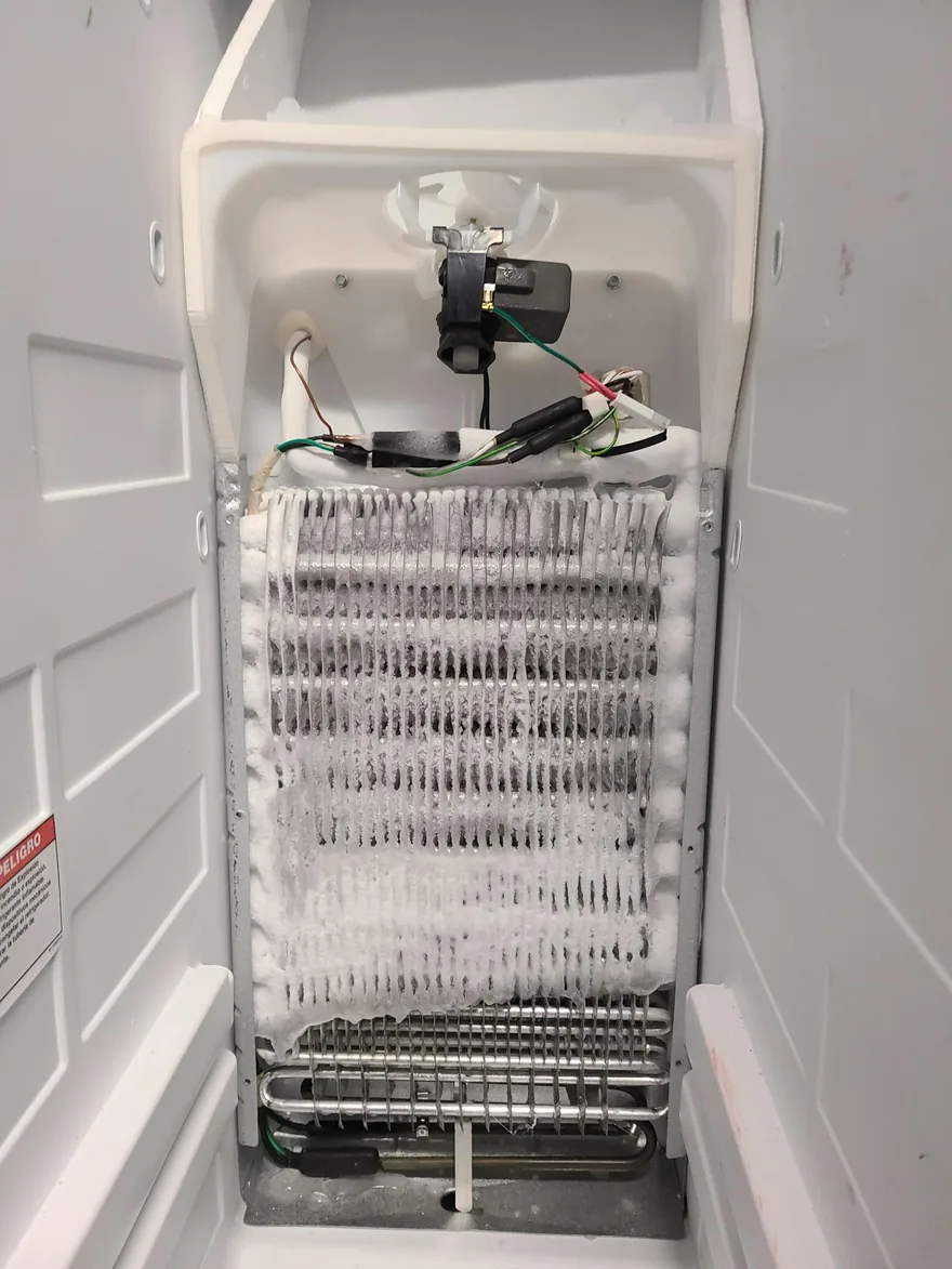 A refrigerator evaporator coil completely iced over behind the freezer panel, with the fan motor and defrost wiring exposed